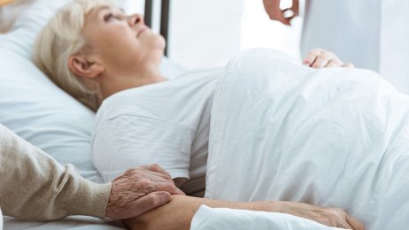 Syk kvinne Bildet viser en eldre kvinne som ligger i en seng og en mann som holder henne i armen