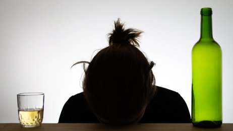 Bildet viser silhuetten av en kvinne som hviler hodet på en bordkant. Ved siden av henne står et glass med alkohol og en flaske.