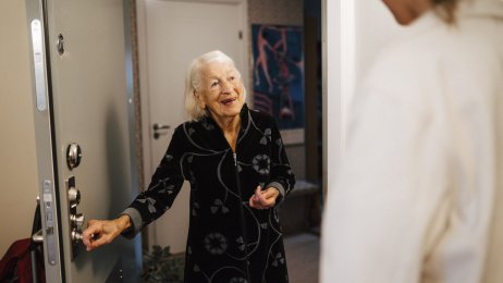 Hjemmesykepleie Bildet viser en smilende eldre kvinne som åpner døren for hjemmesykepleien