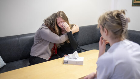 Bildet viser en ung, gråtende pasient som får en klem av pårørende. Bildet viser også en sykepleier som sitter på motsatt side av bordet.