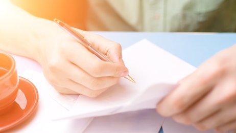 Bildet viser et utsnitt av et menneske som sitter og skriver. Vedkommende har en tekopp ved siden av seg, og solen skinner inn i værelset. Bildet viser et utsnitt av et menneske som sitter og skriver. Vedkommende har en tekopp ved siden av seg, og solen skinner inn i værelset.