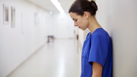 Kvinnelig helsearbeider Kvinnelig helsearbeider står inntil veggen i en sykehuskorridor og kikker i golvet. Hun virker sliten eller fortvilet.