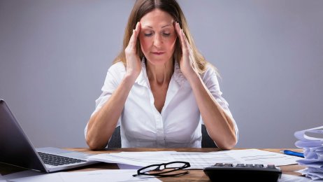 Sliten leder Bildet viser en kvinne som sitter foran et skrivebord med forskjellige permer og rekvisita på og holder seg til tinningene