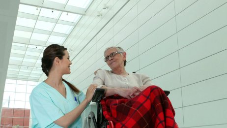 Pleier og dame i rullestol Bildet viser en kvinnelig pleier som sitter på huk ved siden av en eldre dame i rullestol