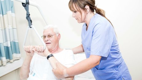 Sykepleier Bildet viser en sykepleier som hjelper en eldre mann med å sette seg opp i sengen