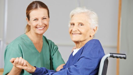 Sykepleier og pasient Bildet viser en sykepleier som holder en eldre pasient i hånda. Pasienten sitter i rullestol, og begge smiler.