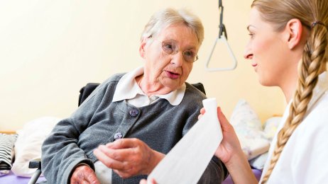 God kommunikasjon mellom sykepleier og pasient Bildet viser en eldre dame hjemme hos seg selv, og en sykepleier som ruller ut en bandasje mens hun snakker med pasienten.