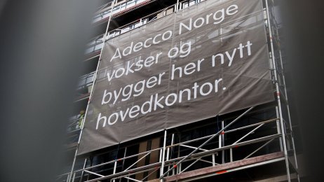 Bildet viser fasaden av et bygg under oppføring, med et banner der det står Adecco Norge vokser og bygger her nytt hovedkontor