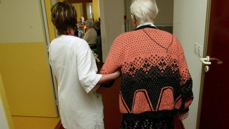 På sykehjem En pleier leier en sykehjemspasient mot spisestuen. Bildet er tatt bakfra.