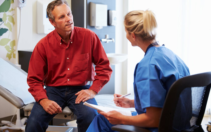 Bildet viser en mannlig pasient som er i konsultasjon med en kvinnelig sykepleier. Han sitter på en sykehusseng, og hun har et skjema i hånda.