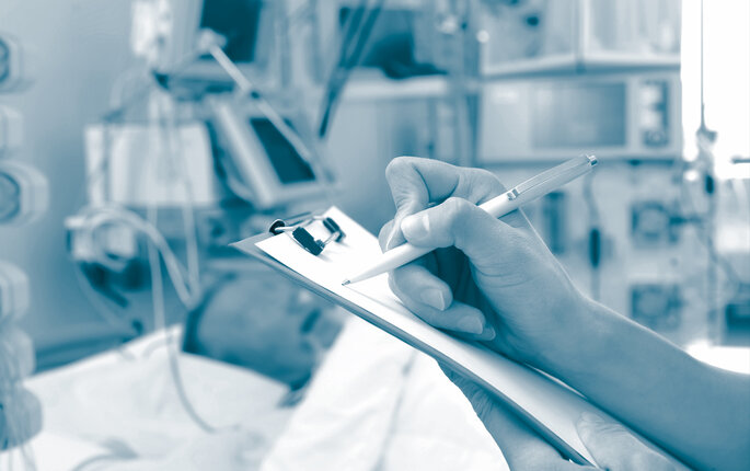 The photo shows an ill patient in an ICU. In the foreground a nurse's hands are holding a form while writing on the form