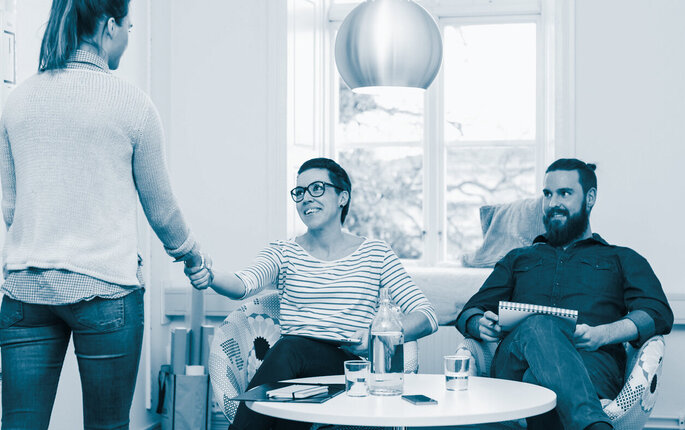 The photo shows a young woman standing with her back towards the reader. She is shaking hands with a smiling woman sitting by a small table. Beside her there is a smiling man. There are books and folders in the bookcase up against the wall..