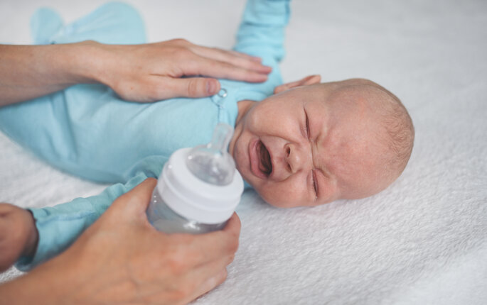 The photo shows a crying infant. A soothing hand lies comforting on the baby's stomach. The other hand is holding a bottle ready to feed the baby