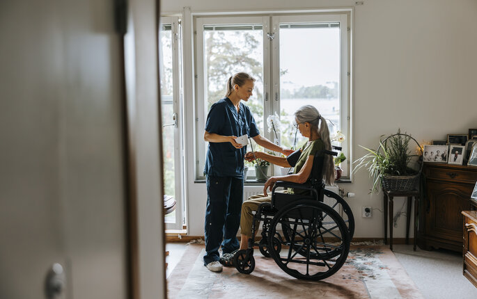 The phowo shows a nurse standing in front of a woman in a wheel chair. The nurse is monitoring her blood pressure. The are in front of a window in the patient's living room