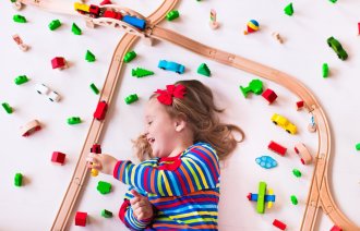Jente i førskolealdre med krøllete hår ligger på ryggen på gulvet midt i en togbane av tre. Leker og smiler.