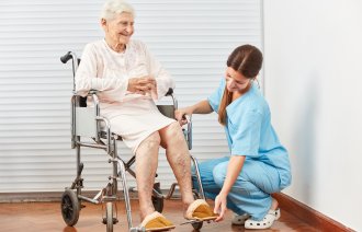 Bildet viser en sykepleier som sitter på golvet og hjelper en eldre dame i en rullestol.