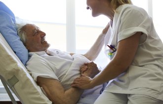 Bildet viser en pasient som ligger i senga, og sykepleieren sitter på sengekanten og holder ham på armen. Begge ler.