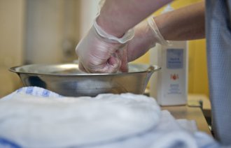 Bildet viser to hender med hansker som vrir opp en klut i et vaskevannsfat.