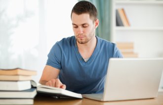 Bildet viser en mannlig student som leser i bøker og skriver på en laptop
