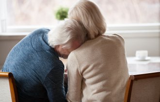 Bildet viser et eldre par som sitter inntil et bord. Mannen lener hodet sitt på kvinnens skulder.