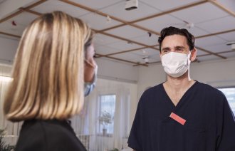 Mann og kvinne iført ansiktsmasker Bildet viser en mann og en kvinne, begge iført ansiktsmasker, som snakker sammen