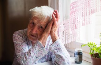Bildet viser en trist, gammel dame som lener hodet mot hendene sine. Hun sitter ved kjøkkenbordet med et pilleglass foran seg.