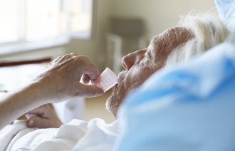 Bildet viser en eldre, syk mann i senga som drikker medisin fra et plastglass