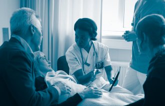 Bildet viser en sykepleier som forklarer noe til en pasient i senga og pasientens pårørende som står rundt