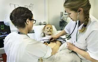 Bildet viser to sykepleiere som står ved en seng. I senga ligger en simuleringsdukke. Den ene sykepleieren lytter etter hjerteslag på dukken