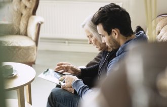 Bildet viser en eldre dame på sykehjem som ser på et nettbrett sammen med en pleier