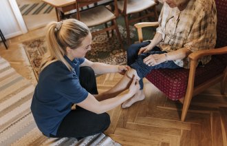 Bildet viser en hjemmesykepleier som skifter strømper på en eldre dame som sitter i stolen i hjemmet sitt.