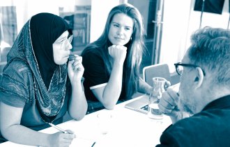 Meeting The photo shows a woman wearing a hijab, a woman wearing Western clothes and a man in a suit, sitting around a table. They are attending a meeting.