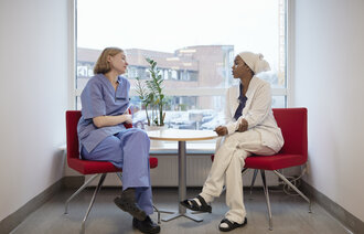 The photo shows two nurses sitting and talking together in front of a window