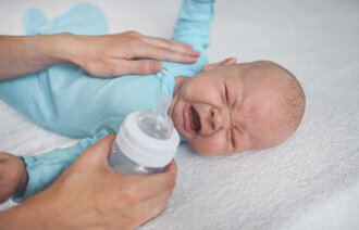 The photo shows a crying infant. A soothing hand lies comforting on the baby's stomach. The other hand is holding a bottle ready to feed the baby