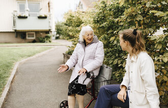 Bildet viser en smilende kvinne med rullator som snakker med en pleier ute
