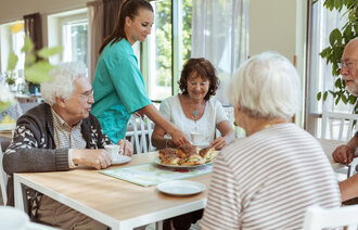 Bildet viser fire eldre som sitter rundt et bord. En sykepleier serverer kanelboller til dem