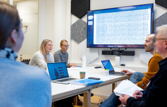 The photo shows two students sitting around a table opposite two academic supervisors. A third student can be seen with her back towards the camera. At the screen on the wall graphs of cardiac arrhythmias can be seen. The people are participating in a reflection group.