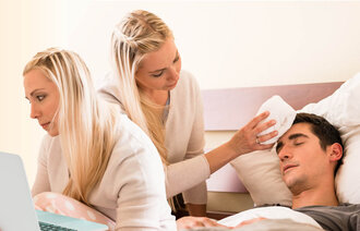 Partner of someone with cancer The photo shows a young woman working in front of a laptop. The same woman can be seen to the right in the photo, where she is wiping an ill man’s forehead.