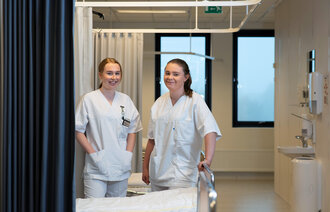 Bildet viser to sykepleiere på sengepost som ser mot kamera