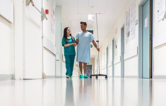 Bildet viser en sykepleier og en pasient som går i sykehuskorridoren sammen