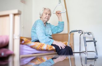 Gammel dame på pleiehjem sitter på sengen med rullator foran. Rutete pledd.