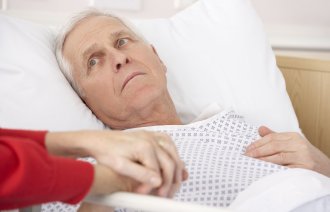 Bildet viser en syk, eldre mann som ligger i en sykehusseng. En dames hender holder hans.