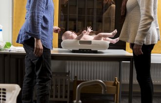 Bildet viser en mor som er på helsestasjonen, der babyen veies av helsesøster.