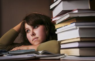 Bildet viser en student med et litt oppgitt uttrykk, som ligger over en oppslått bok på pulten. Det ligger en stabel bøker ved siden av henne.