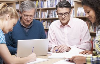 Bildet viser en gruppe mennesker som sitter rundt et bord og arbeider med papirer. Bak er det bokhyller.