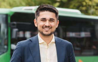 Portett av ung mann med indisk bakgrunn, smilende i dress foran grønn buss