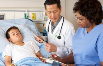 Bildet viser et barn i sengen på barneavdeling, og legen og sykepleieren står ved siden av og noterer på skjemaer.