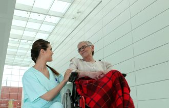 Bildet viser en kvinnelig pleier som sitter på huk ved siden av en eldre dame i rullestol