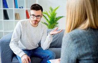 Bildet viser en ung mann som sitter i en sofa, tvers overfor en kvinnelig terapeut, som vi ser ryggen av.