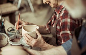 Bildet viser en eldre kvinne og en eldre mann som lager og dekorerer gjenstander i leire